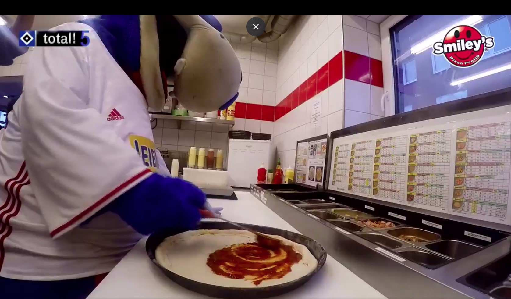 Dino Hermann, das Maskottchen vom HSV in einer Smiley's Pizza einen Pizza Teil belegend stehend