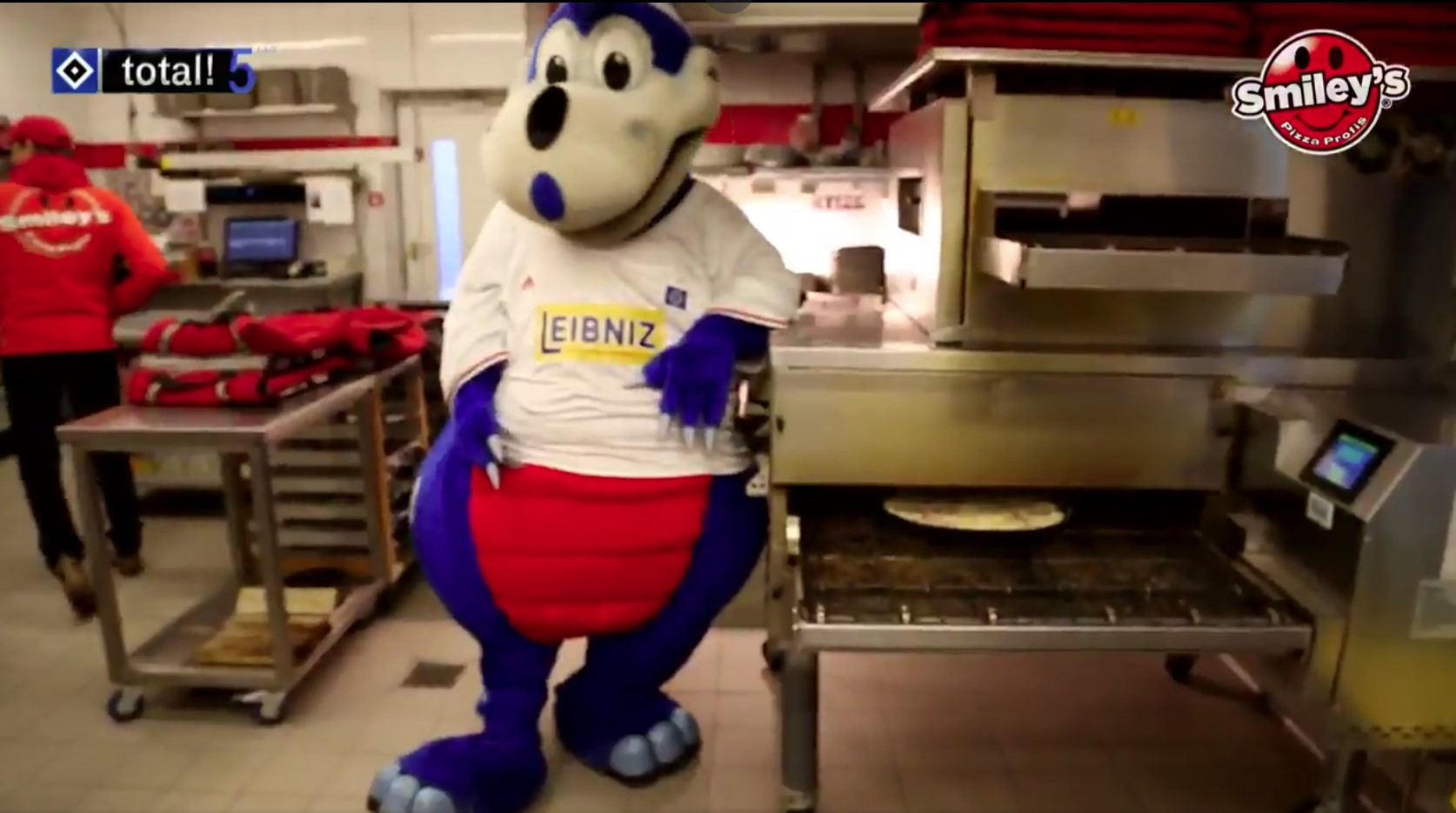 Dino Hermann, das Maskottchen vom HSV in einer Smiley's Pizza Filiale am Pizza-Ofen stehend